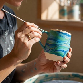 Someone with a paint pallet with a paint brush about to paint a pottery mug